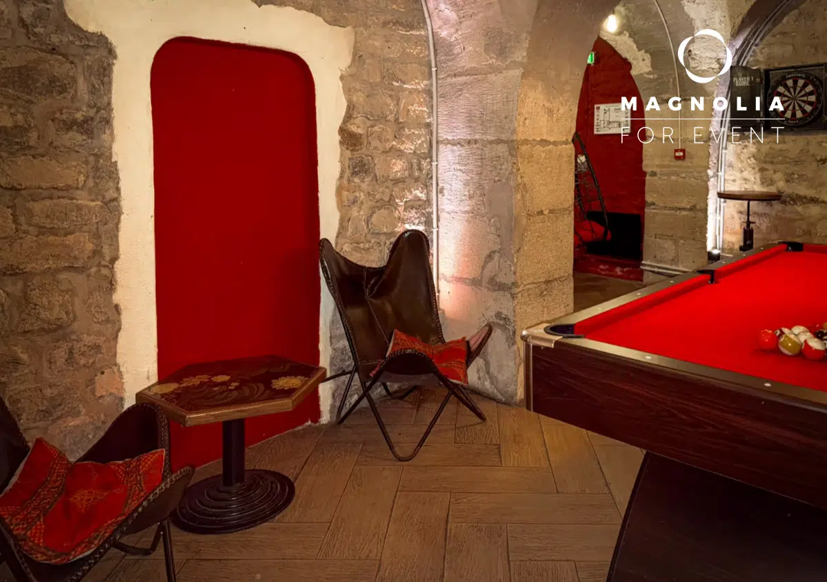 Un salon confortable au PETIT PARIS CACHÉ (75004) avec des murs en pierre, deux chaises en cuir, une table ronde et une porte rouge. Une partie d'une table de billard est visible sur la droite ; "Magnolia For Event" apparaît dans le coin supérieur droit.