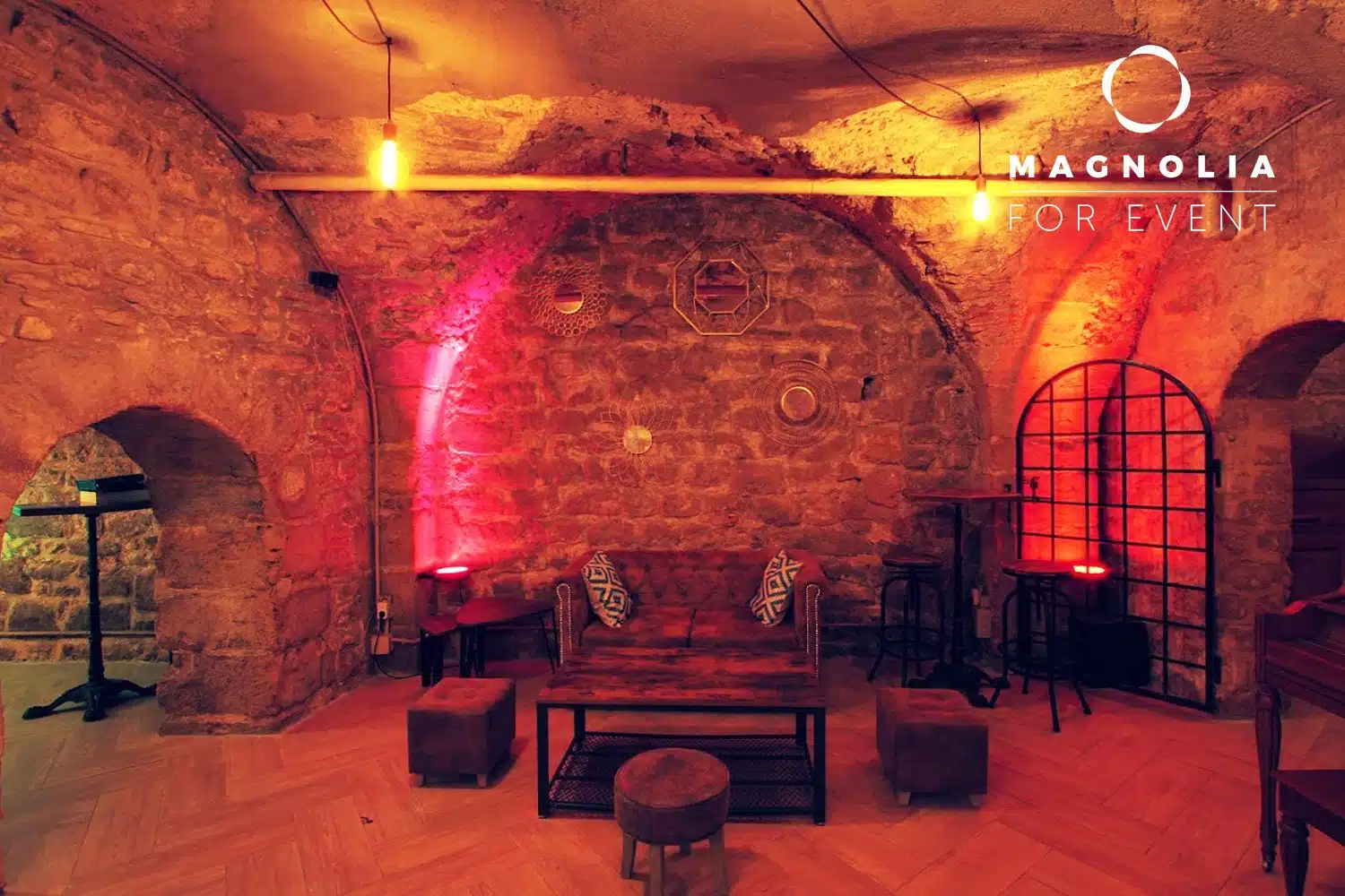 Charmant espace événementiel avec des murs en pierre, un éclairage chaleureux, un mobilier rustique, des coussins et des œuvres d'art murales géométriques. Le logo "LE PETIT PARIS CACHÉ (75004)" apparaît dans le coin supérieur droit.
