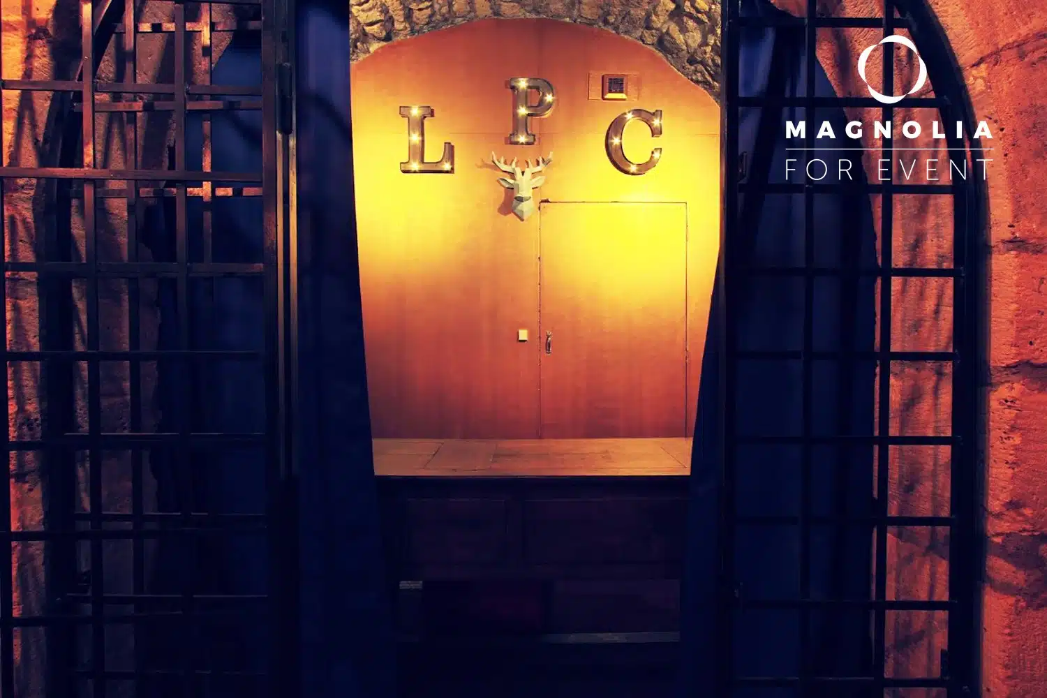Une entrée faiblement éclairée avec des grilles en métal noir mène à un mur en bois affichant de grandes lettres métalliques "LPC" et une tête de cerf. La mention "Magnolia For Event" est visible en haut à droite. Produit : LE PETIT PARIS CACHÉ (75004).