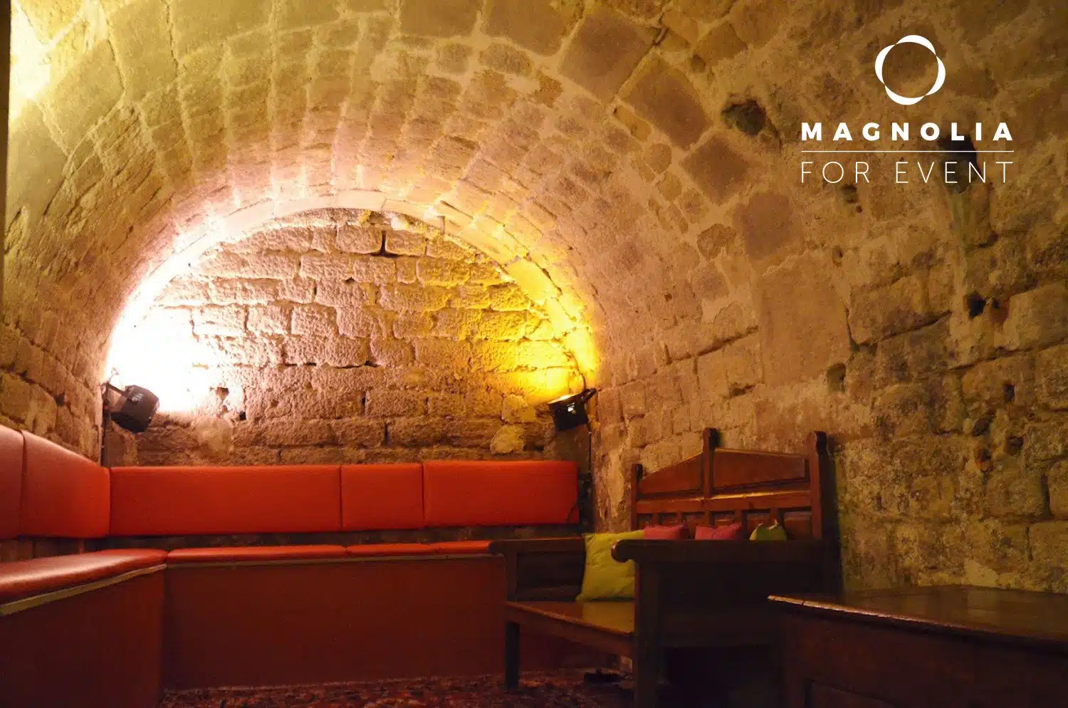 Une salle souterraine confortable aux murs de pierre avec un plafond voûté, un éclairage jaune chaud, des banquettes rouges et des meubles en bois au PETIT PARIS CACHÉ (75004). Le logo de Magnolia For Event se trouve dans le coin supérieur droit.