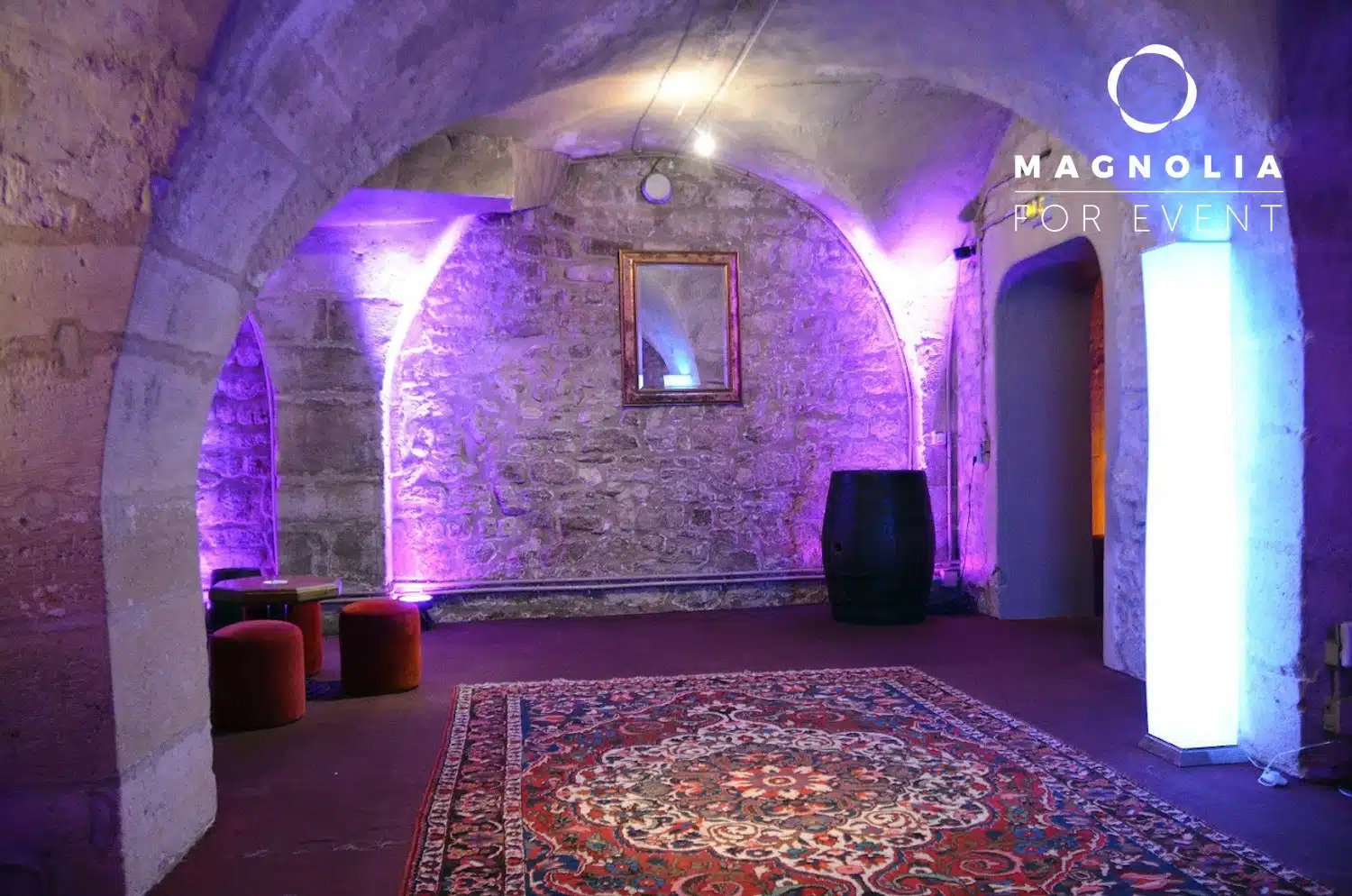 Une pièce confortable aux murs de pierre avec un éclairage violet, un tapis orné, un tonneau, des tabourets rouges et un miroir encadré. Le logo Magnolia For Event apparaît dans le coin supérieur droit. Lieu : LE PETIT PARIS CACHÉ (75004).