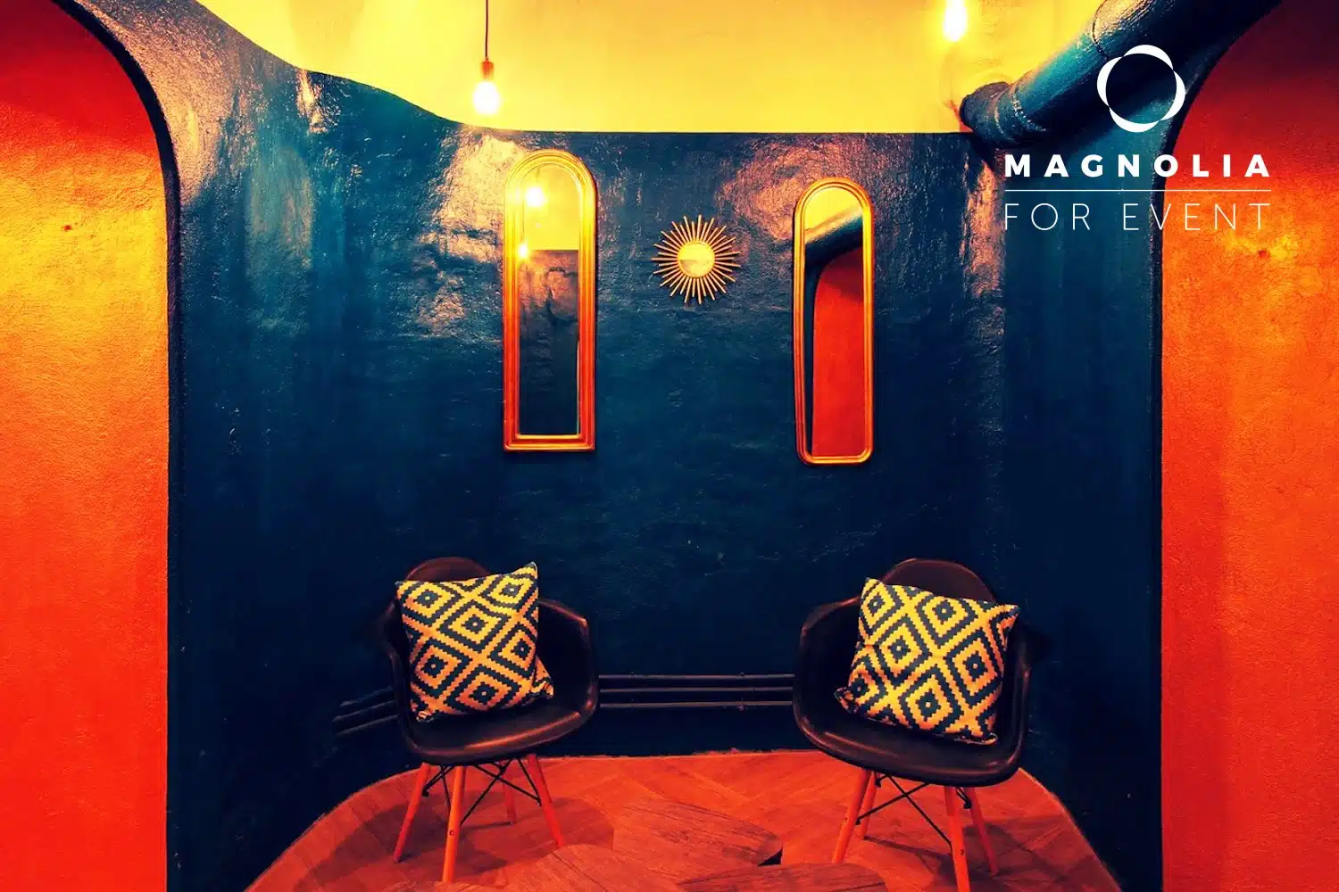 Deux chaises noires avec des coussins à motifs se font face dans une pièce confortable aux murs bleu foncé, aux miroirs encadrés d'or, à la décoration murale en forme de soleil et à l'éclairage chaleureux. Le mur porte l'inscription "LE PETIT PARIS CACHÉ (75004)".