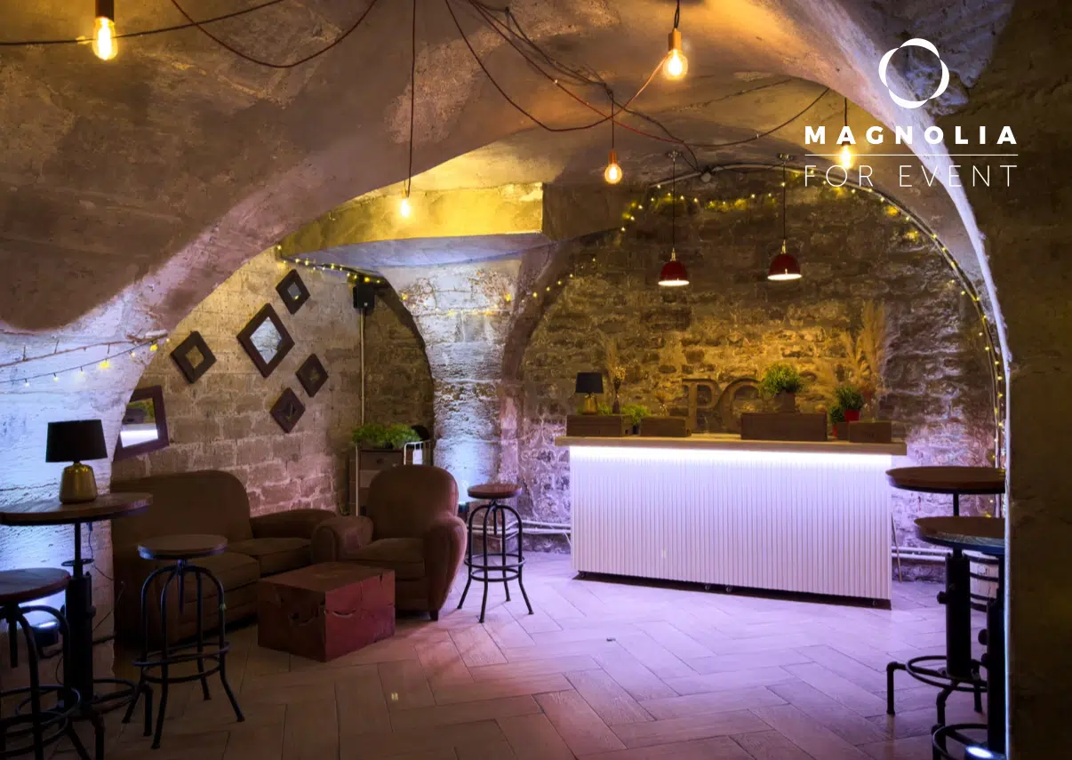 Un espace confortable et rustique avec des murs en pierre et des plafonds voûtés, avec un bar lumineux, des fauteuils bruns, des petites tables, des lampes suspendues et des plantes en pot ; le logo "LE PETIT PARIS CACHÉ (75004)" apparaît dans le coin supérieur droit.