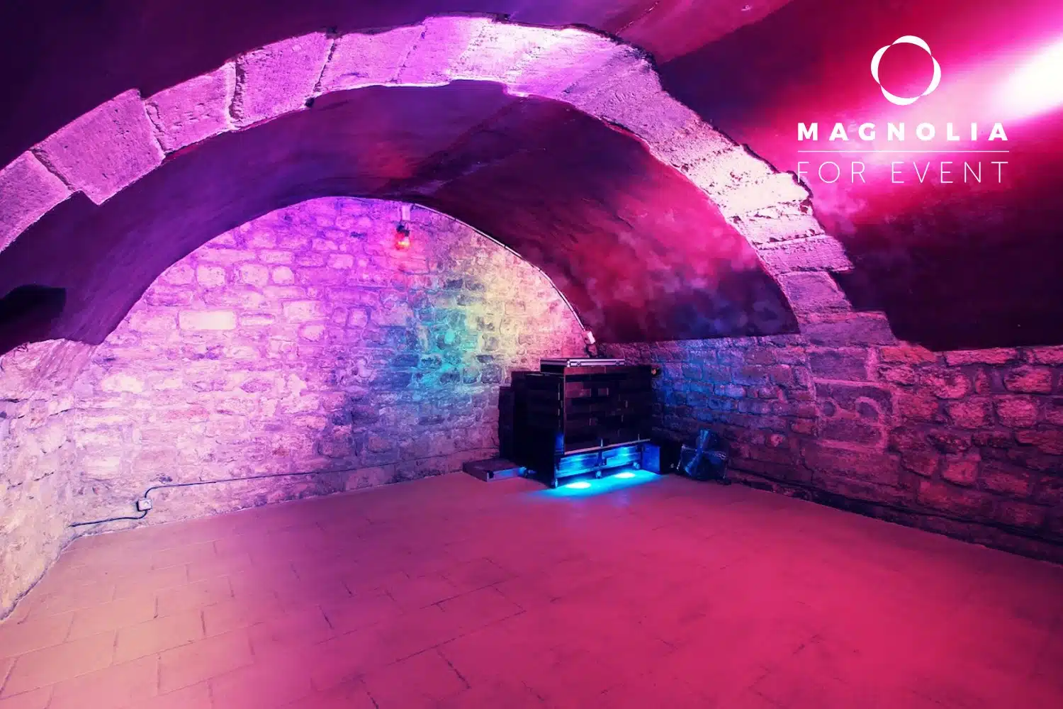 Sous-sol aux murs de pierre et au plafond voûté du PETIT PARIS CACHÉ (75004) avec un éclairage violet et bleu, un sol carrelé et une console d'équipement noire dans le coin. Le logo de Magnolia for Event apparaît en haut à droite.