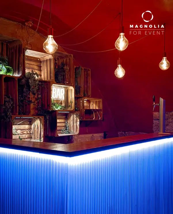 LE PETIT PARIS CACHÉ (75004) est un bar moderne avec un comptoir bleu lumineux, des étagères en bois avec des plantes, des ampoules apparentes au plafond rouge et une enseigne BAR en néon. Le logo Magnolia For Event apparaît dans le coin supérieur droit.