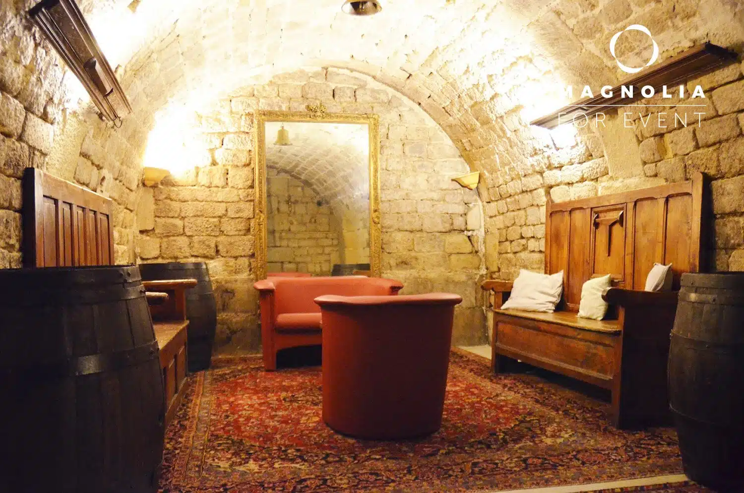 LE PETIT PARIS CACHÉ (75004) : Une cave confortable aux murs de pierre avec un plafond voûté, un grand miroir, des banquettes rembourrées, deux fauteuils rouges, une table ronde, un décor de tonneau, un tapis à motifs et un éclairage chaleureux pour une atmosphère accueillante.