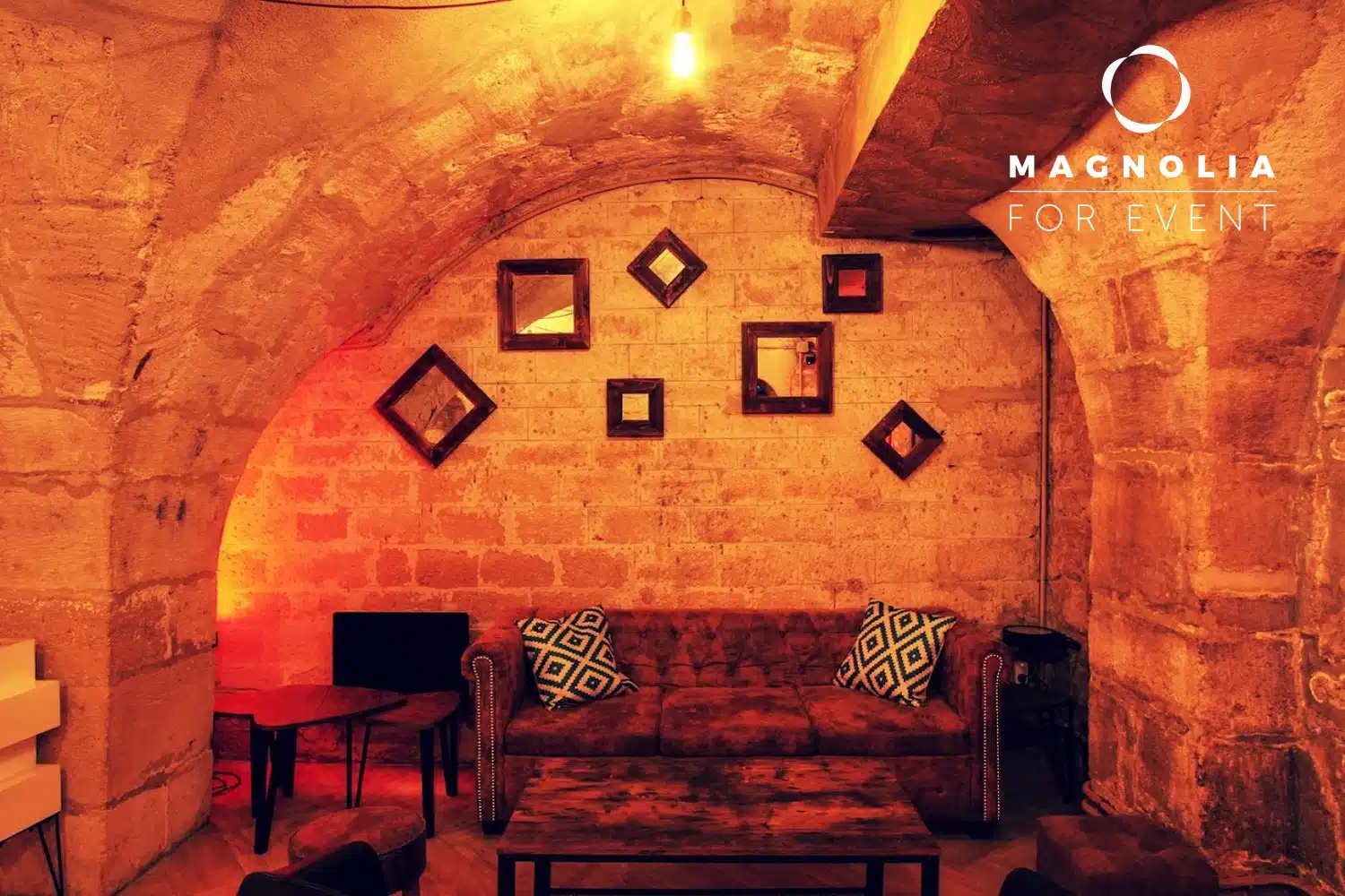 Un salon confortable, faiblement éclairé, avec un plafond en pierre incurvé, des murs en briques apparentes, un canapé marron avec des coussins à motifs, des petites tables et des miroirs décoratifs. La mention "LE PETIT PARIS CACHÉ (75004)" apparaît dans le coin supérieur droit.