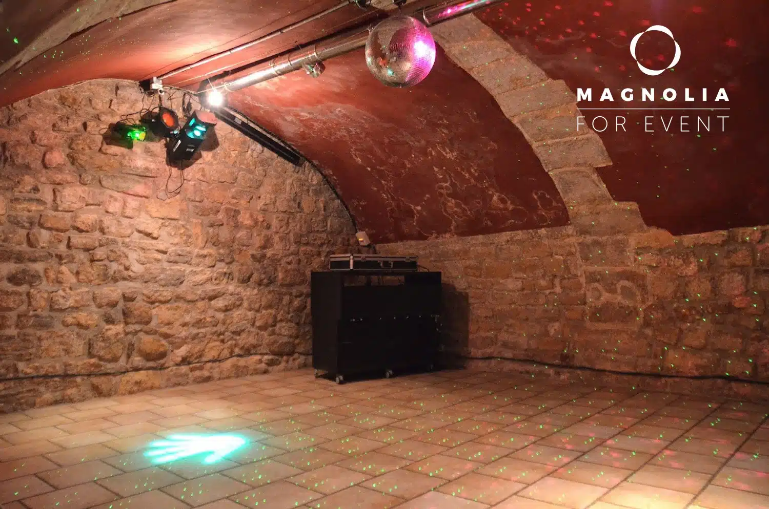 LE PETIT PARIS CACHÉ (75004) : Un espace rustique, aux murs de pierre, avec un sol carrelé, un plafond voûté, une boule à facettes, des lumières colorées et du matériel de sonorisation ; le logo de Magnolia For Event dans le coin supérieur droit.