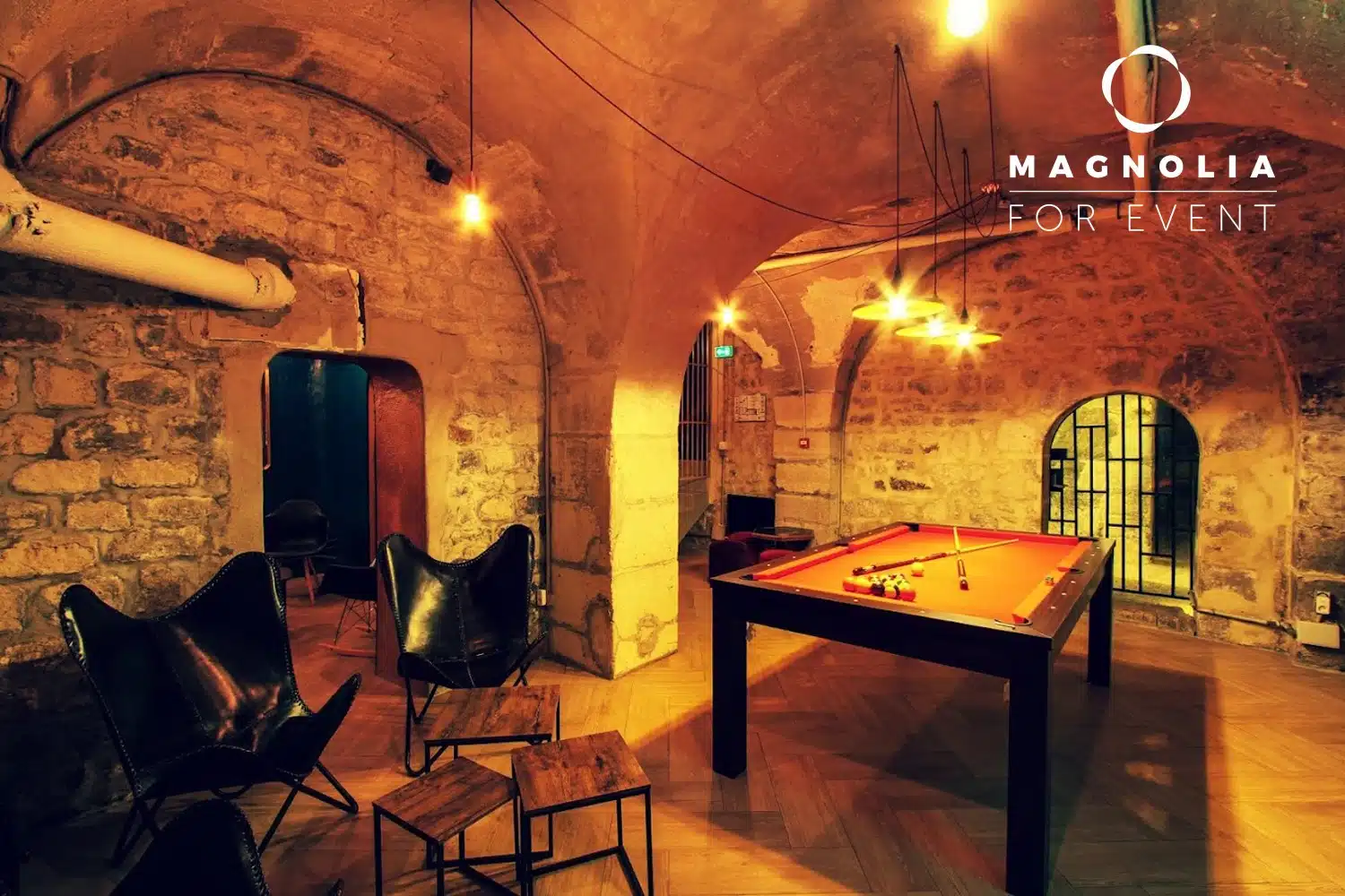 Une pièce confortable et faiblement éclairée, située dans une cave en pierre au plafond voûté, est équipée d'une table de billard rouge, de chaises en cuir noir et d'ampoules électriques suspendues. Le logo LE PETIT PARIS CACHÉ (75004) apparaît dans le coin supérieur droit.