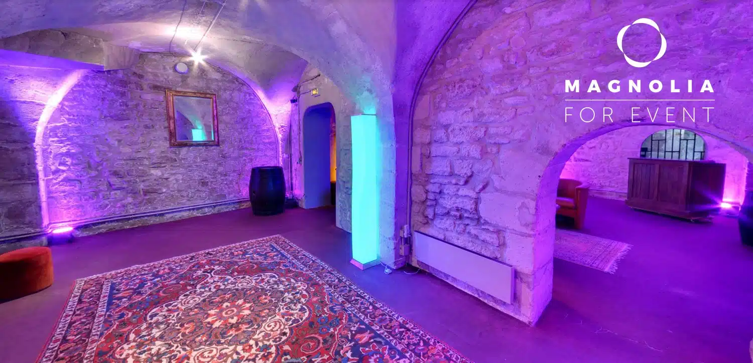 LE PETIT PARIS CACHÉ (75004) est un espace événementiel aux murs de pierre avec des portes cintrées, un éclairage violet, un tapis à motifs et un décor moderne. Le nom du lieu et un logo circulaire sont apposés sur le mur de droite.