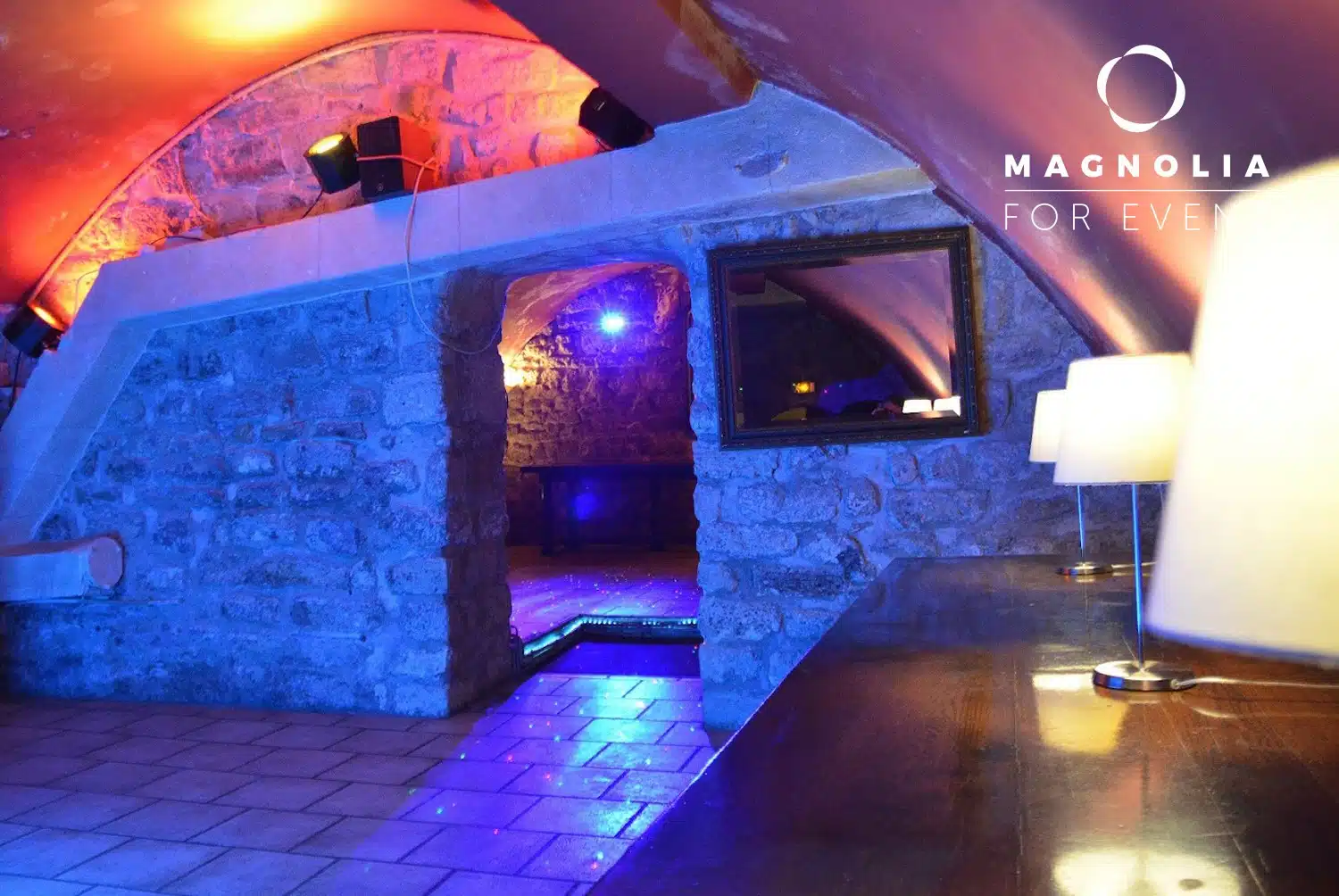 LE PETIT PARIS CACHÉ (75004) est un espace confortable, faiblement éclairé, avec des murs en pierre, des plafonds voûtés, des lampes modernes et un éclairage d'appoint bleu-orange. Le logo Magnolia For Event apparaît dans le coin supérieur droit.