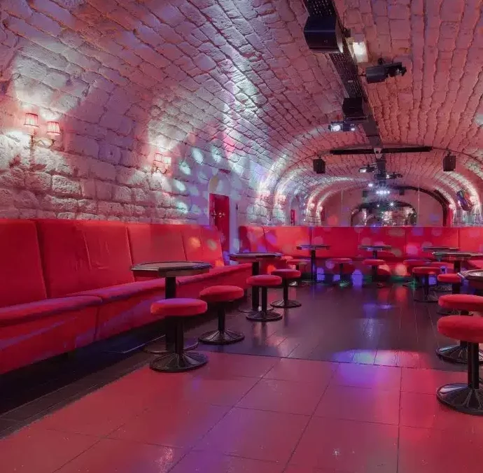 LE LIEU SECRET (75005) est une boîte de nuit moderne avec des plafonds voûtés en pierre, des sièges rouges, des tables rondes et des lumières colorées qui se reflètent sur le sol et les murs, créant une atmosphère élégante et confortable dans un espace vide.