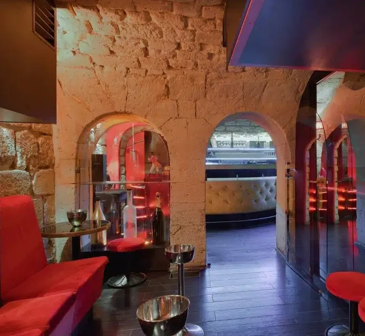 LE LIEU SECRET (75005) se caractérise par des arcades en pierre, des sièges rouges rembourrés, de petites tables rondes et un éclairage tamisé. Un comptoir de bar moderne et des miroirs ajoutent une touche élégante à ce lieu historique.