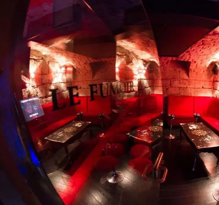 Salon faiblement éclairé avec des sièges en velours rouge, des tables en bois sombre, des murs en pierre et des appliques chaleureuses. La mention "LE LIEU SECRET (75005)" brille sur le verre, ce qui correspond parfaitement à l'ambiance intime et confortable de la pièce.