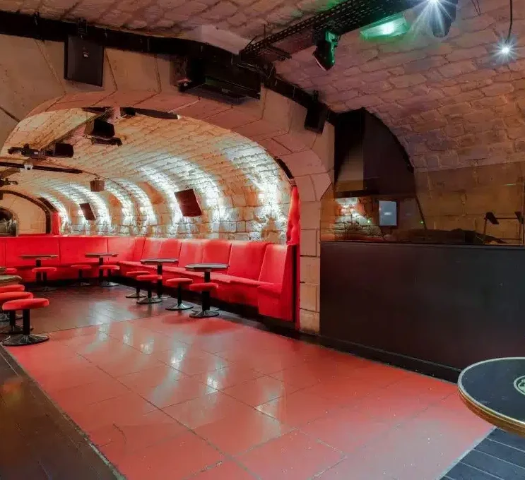 LE LIEU SECRET (75005) est un salon souterrain chic avec des plafonds voûtés en pierre, des sols en carreaux rouges et des banquettes coussinées le long du mur, doucement éclairées par des spots qui accentuent la pierre.