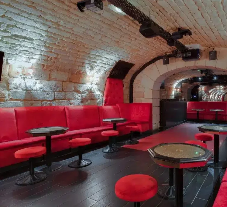 LE LIEU SECRET (75005) est un salon élégant avec des plafonds voûtés en pierre, des banquettes rouges rembourrées, des tables rondes et des tabourets. L'éclairage moderne et les planchers en bois foncé créent une atmosphère confortable et haut de gamme.
