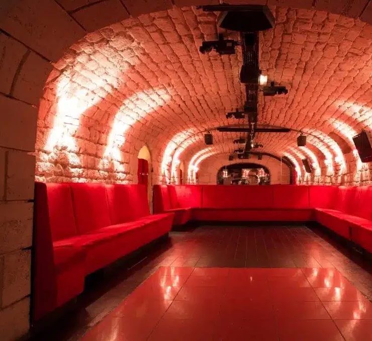 LE LIEU SECRET (75005) dispose d'un salon confortable aux murs de pierre voûtés, avec des banquettes rouges rembourrées de chaque côté. Un éclairage chaleureux met en valeur le plafond de briques, créant ainsi une atmosphère accueillante et conviviale.