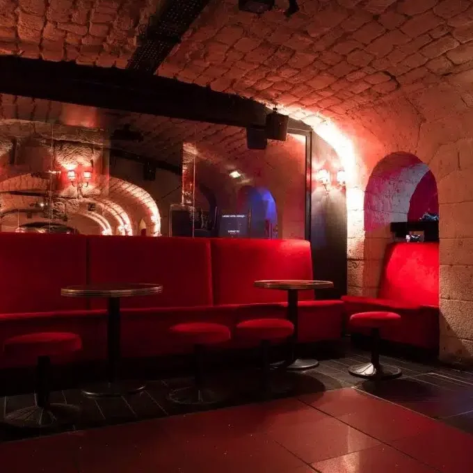 LE LIEU SECRET (75005) propose un salon confortable avec des banquettes en velours rouge, des tabourets ronds, des plafonds en pierre voûtés et un éclairage chaleureux qui brille sur les murs en miroir dans un bar faiblement éclairé.
