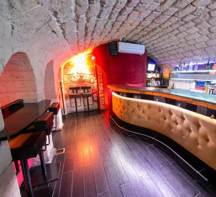 LE LIEU SECRET (75005) dispose d'un bar confortable avec un comptoir incurvé et touffeté, un plafond en pierre, un éclairage moderne, des tables hautes à gauche et des étagères de verres et de bouteilles derrière le bar.