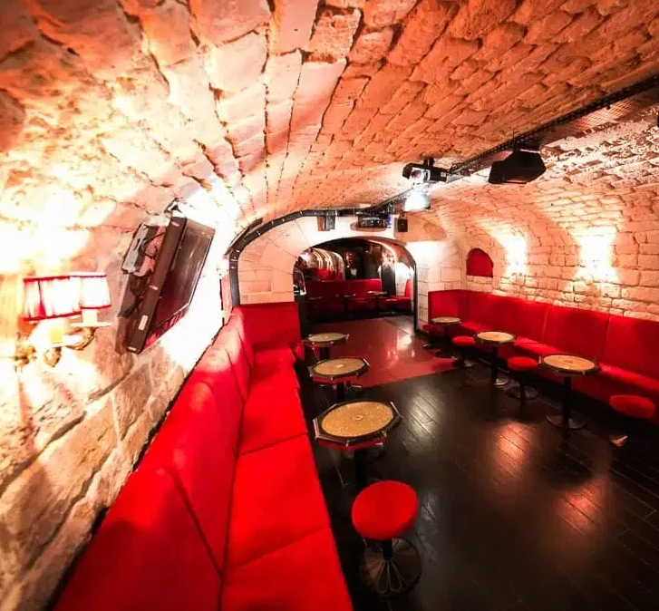 LE LIEU SECRET (75005) est un salon souterrain confortable et voûté avec des plafonds en briques apparentes, des banquettes aux coussins rouges, des petites tables rondes et un éclairage doux pour une atmosphère chaleureuse et invitante.