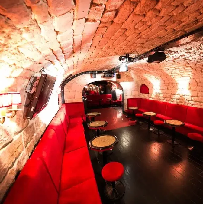 LE LIEU SECRET (75005) est un salon confortable, faiblement éclairé, avec des plafonds voûtés en briques, des sièges rouges rembourrés, des petites tables rondes et un sol sombre et poli pour une ambiance intime et souterraine.