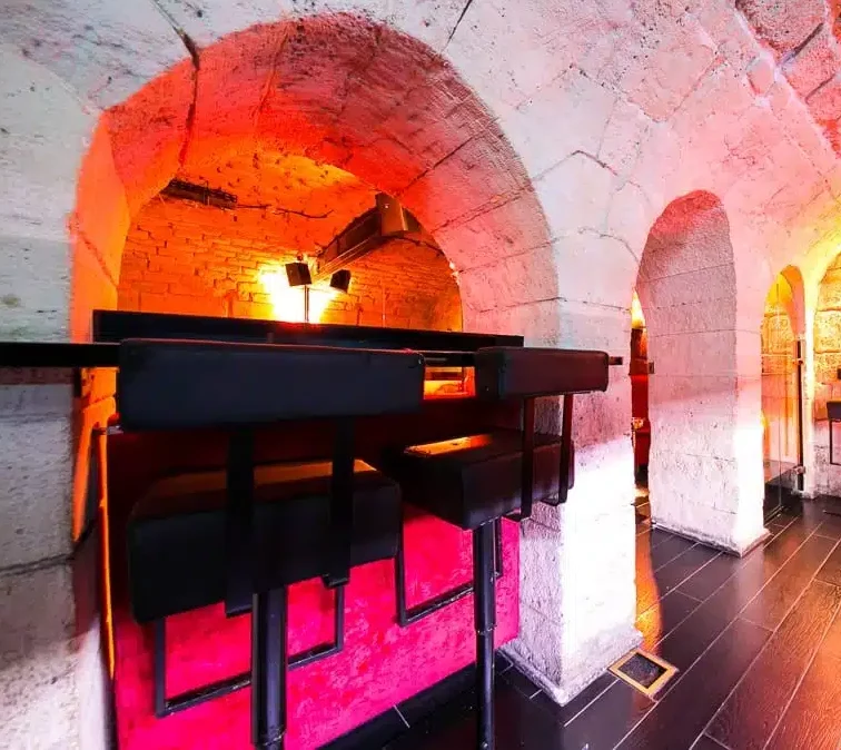 LE LIEU SECRET (75005) présente des murs en pierre, des alcôves voûtées, des chaises noires élégantes, des lumières orange brillantes et des sols sombres polis pour une ambiance souterraine confortable et élégante.