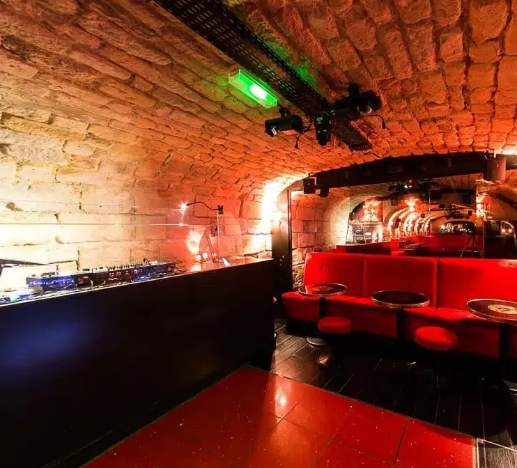 LE LIEU SECRET (75005) est une boîte de nuit confortable avec des plafonds en pierre voûtés, des cabines aux coussins rouges, des petites tables rondes, un éclairage tamisé et une cabine de DJ, créant une ambiance chaleureuse et intime.