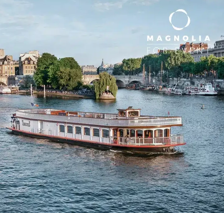 Un bateau fluvial portant l'inscription LE NASHVILLE TENNESSE (75015) navigue sur un fleuve calme, encadré par des arbres, des bâtiments historiques et une petite île avec un saule, sous un ciel partiellement nuageux.