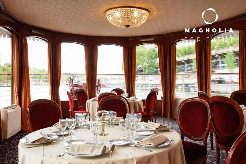 Salle à manger élégante au LE NASHVILLE TENNESSE (75015) avec des tables rondes, de la verrerie, des nappes blanches, des chaises rouges, de grandes fenêtres avec des rideaux bruns, un lustre, et le logo Magnolia For Event dans le coin.