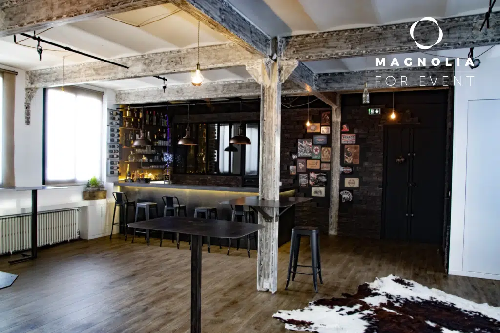 LE JAZZ HARMONY (75011) offre un espace événementiel moderne avec des planchers en bois, des poutres apparentes, un bar élégant garni de bouteilles, des tables hautes, un tapis en peau de vache et un éclairage industriel. Le logo Magnolia For Event figure dans le coin supérieur droit.