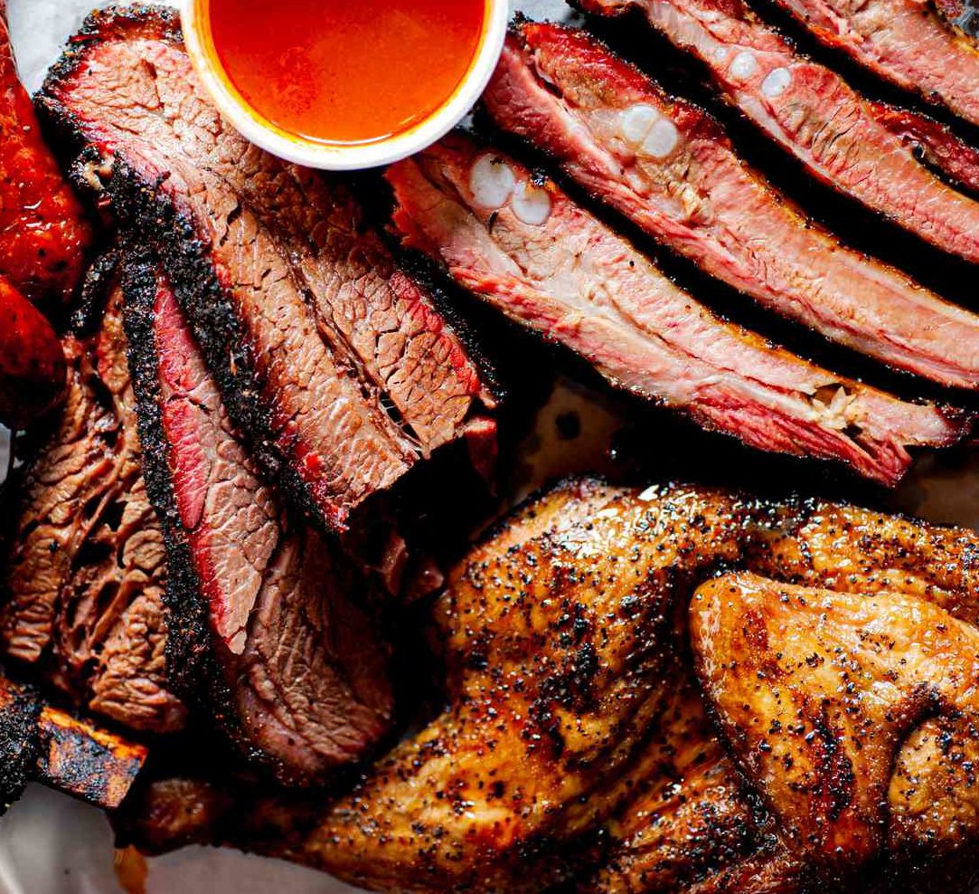 Gros plan sur LE BRISKET SIGNATURE PAR STEPHAN "L'art du feu et du temps", qui présente de la poitrine de bœuf, des côtes, de la saucisse et du poulet assaisonné, carbonisés et juteux, accompagnés d'une sauce barbecue à l'orange.