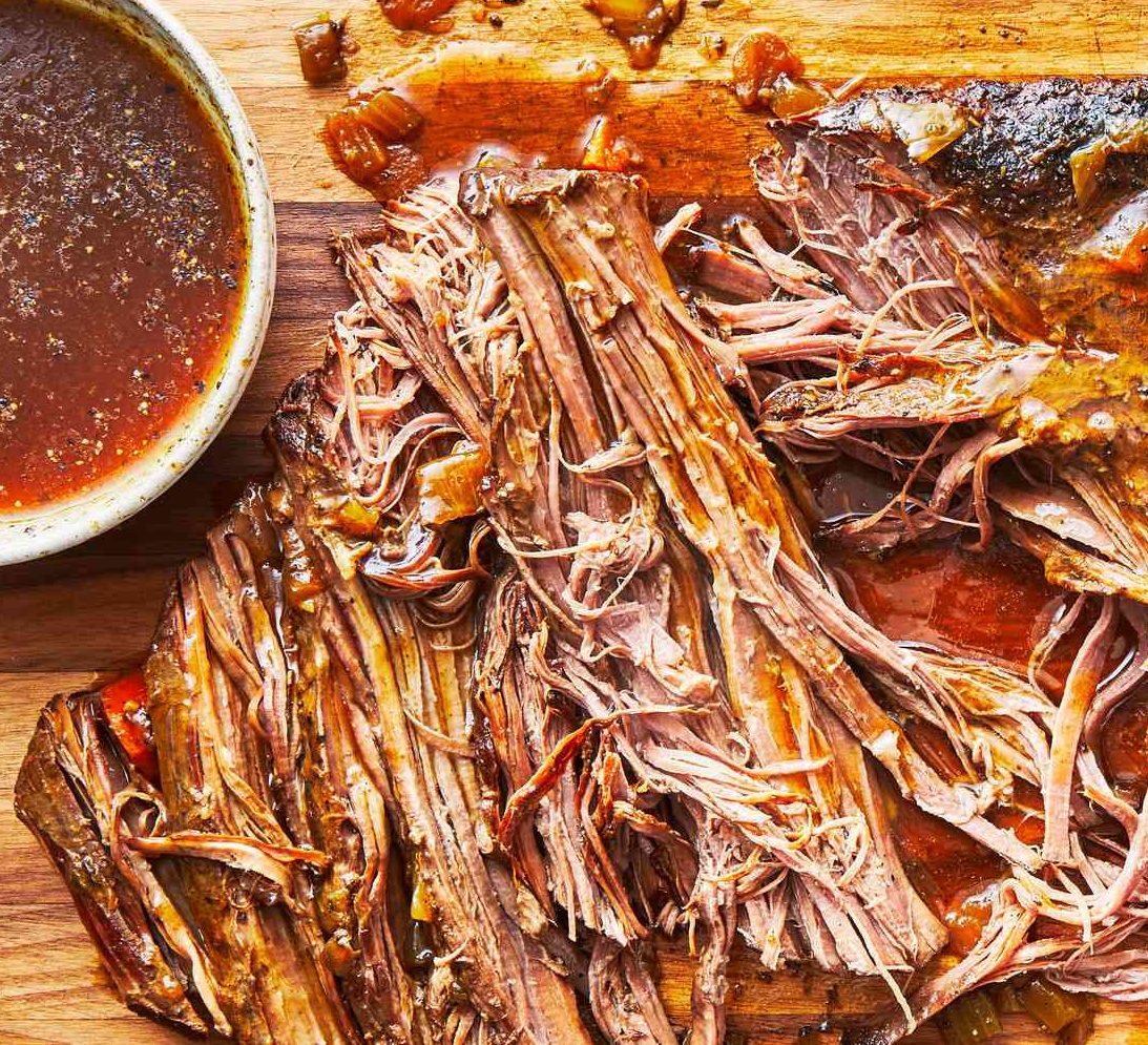 LE BRISKET SIGNATURE PAR STEPHAN "L'art du feu et du temps" présente un rôti de bœuf tendre et juteux, effiloché sur une planche à découper en bois, à côté d'un bol de riche bouillon brun, mettant en valeur sa texture appétissante et sa saveur savoureuse.