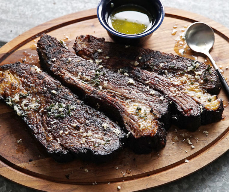 Trois tranches de LE BRISKET SIGNATURE PAR STEPHAN "L'art du feu et du temps", garnies de fromage râpé et d'herbes sur une planche en bois, accompagnées d'un petit bol de sauce verte.