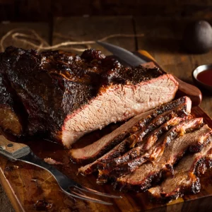 LE BRISKET SIGNATURE PAR STEPHAN "L'art du feu et du temps" est présenté en tranches juteuses sur une planche en bois, révélant sa croûte savoureuse et sa viande tendre, accompagné d'un couteau, d'une fourchette et d'une sauce barbecue en arrière-plan.