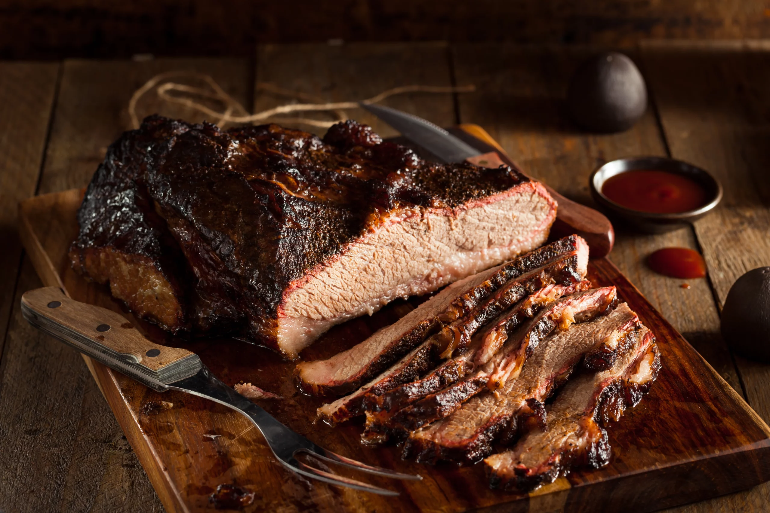 LE BRISKET SIGNATURE PAR STEPHAN "L'art du feu et du temps" est présenté en tranches juteuses sur une planche en bois, révélant sa croûte savoureuse et sa viande tendre, accompagné d'un couteau, d'une fourchette et d'une sauce barbecue en arrière-plan.