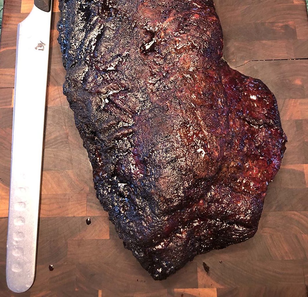 LE BRISKET SIGNATURE PAR STEPHAN "L'art du feu et du temps" - une grosse poitrine de bœuf fumée à la croûte sombre et caramélisée - repose sur une planche à découper en bois à côté d'un long couteau à trancher.
