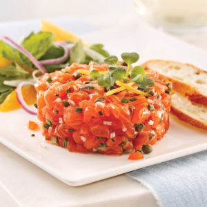 Le TARTARE DE SAUMON propose un tartare de saumon avec des microgreens et un zeste de citron, accompagné de deux tranches de baguette et d'une salade fraîche avec des tranches d'orange et de l'oignon rouge sur une assiette carrée blanche.