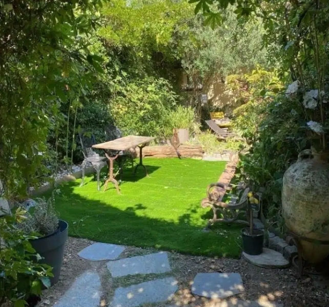 LE LOFT ART FEELING (93170) BAGNOLET présente un jardin luxuriant avec de la verdure vibrante, une petite clairière avec une table et des bancs en bois, des pavés en pierre, et de grandes poteries avec des plantes en pot qui rehaussent l'espace extérieur accueillant.