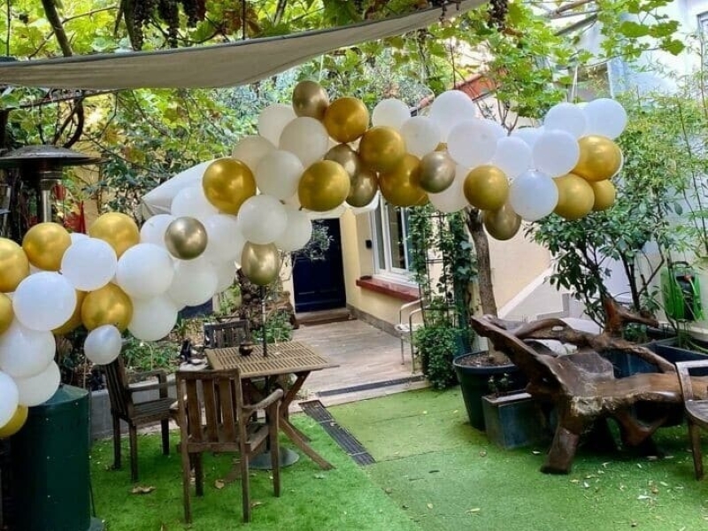 LE LOFT ART FEELING (93170) BAGNOLET : Un jardin avec des ballons blancs et dorés, des tables et des chaises en bois, du gazon artificiel, des plantes en pot et un banc-sculpture en bois près d'une maison de couleur crème.