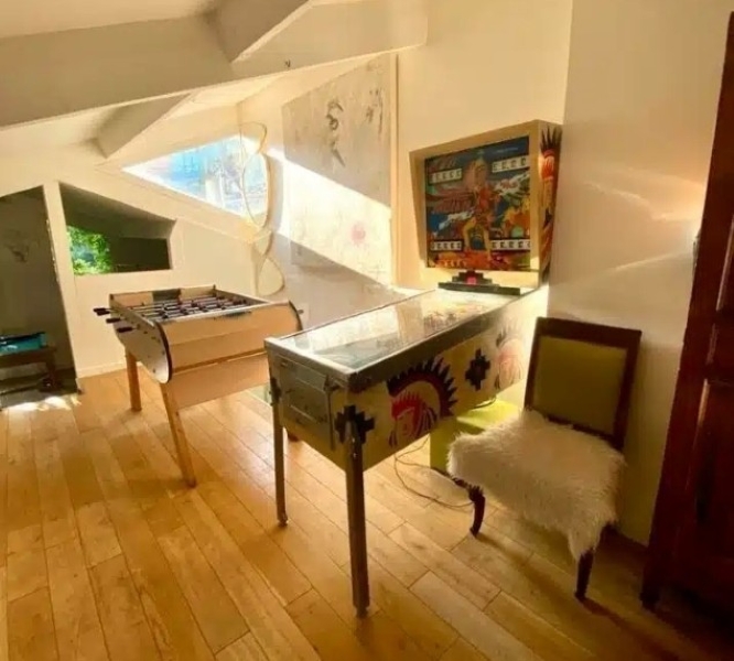 LE LOFT ART FEELING (93170) BAGNOLET dispose d'une salle ensoleillée avec des planchers en bois, d'un baby-foot, d'un flipper vintage avec des œuvres d'art amérindiennes et d'une chaise avec une housse blanche en fourrure.