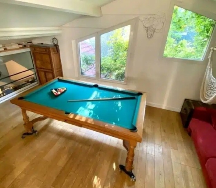 Le BAGNOLET LE LOFT ART FEELING (93170), confortable et lumineux, comprend un petit billard avec des boules et une queue de billard sur un sol en bois près de grandes fenêtres, ainsi qu'un canapé rouge, une chaise hamac et une armoire en bois.