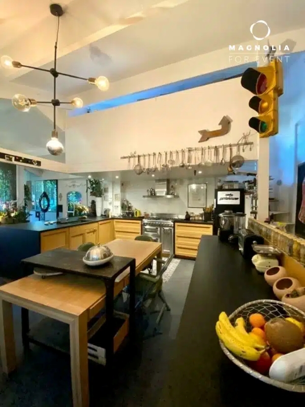 La cuisine moderne de LE LOFT ART FEELING (93170) BAGNOLET comprend des armoires en bois, un îlot central, des appareils en acier inoxydable, des ustensiles suspendus, un comptoir noir avec une coupe à fruits, et un feu de circulation sur le mur de droite sous des lumières vives.