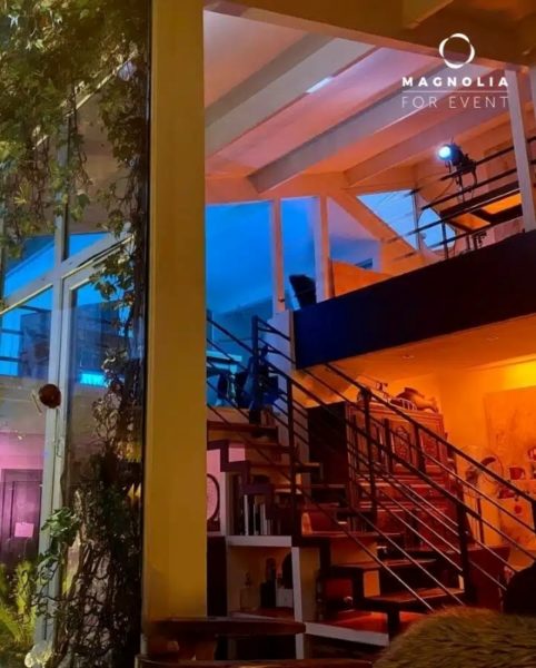Découvrez le BAGNOLET LE LOFT ART FEELING (93170) : un loft moderne et confortable avec un escalier ouvert, des plantes par de grandes fenêtres, un éclairage chaleureux et des teintes bleu-orange. Le logo Magnolia For Event apparaît dans le coin supérieur droit.