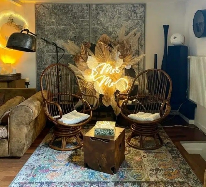 Un salon confortable avec LE LOFT ART FEELING (93170) BAGNOLET, avec deux chaises rondes en osier, des coussins, une table rustique en bois et une enseigne au néon Mrs & Mrs au milieu de feuilles séchées et d'herbe de la pampa pour une ambiance chaleureuse et invitante.