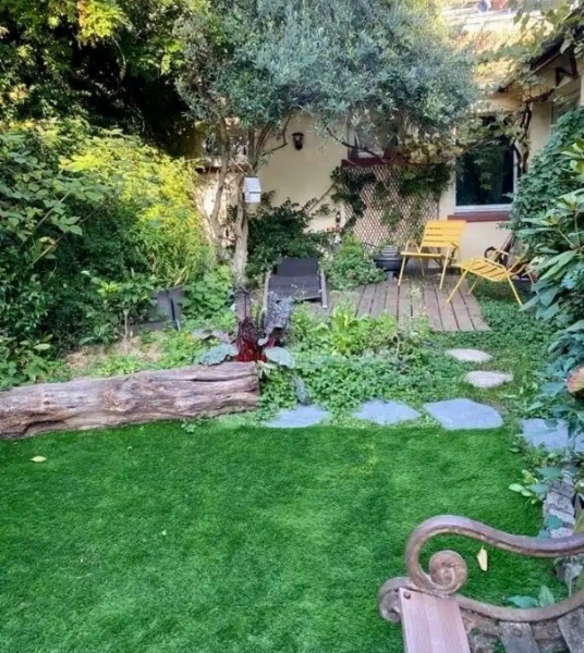Le BAGNOLET LE LOFT ART FEELING (93170) présente un jardin luxuriant avec de l'herbe verte, un banc en bois, un arbre, une bûche et un patio avec des chaises et une table jaunes près de la maison, au milieu de plantes et de verdure éclatantes.