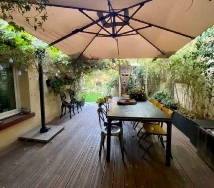 Le BAGNOLET LE LOFT ART FEELING (93170) dispose d'un patio couvert avec un grand parasol, d'une terrasse en bois, d'un ensemble de salle à manger, de bancs sur les côtés sous des plantes suspendues, d'une végétation luxuriante et d'une sculpture décorative en forme de masque à l'arrière.