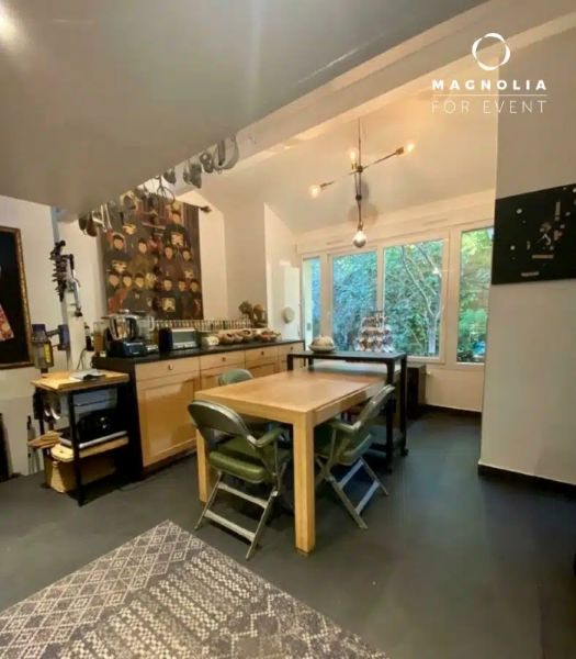 Cuisine et salle à manger modernes dans LE LOFT ART FEELING (93170) BAGNOLET avec une table en bois, quatre chaises, une grande fenêtre avec de la verdure, un luminaire suspendu, des étagères et des plans de travail contenant des appareils électroménagers. Logo MAGNOLIA FOR EVENT dans le coin.