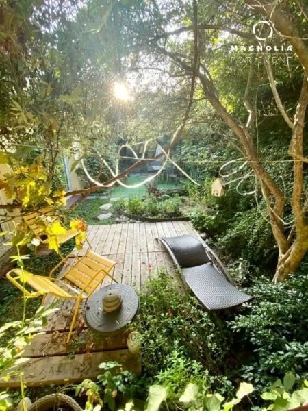 Un jardin ensoleillé avec une terrasse en bois, une chaise en métal jaune, une chaise longue sombre et une végétation luxuriante. La lumière du soleil filtre à travers les arbres, illuminant l'espace. Le logo LE LOFT ART FEELING (93170) BAGNOLET apparaît dans le coin supérieur droit.
