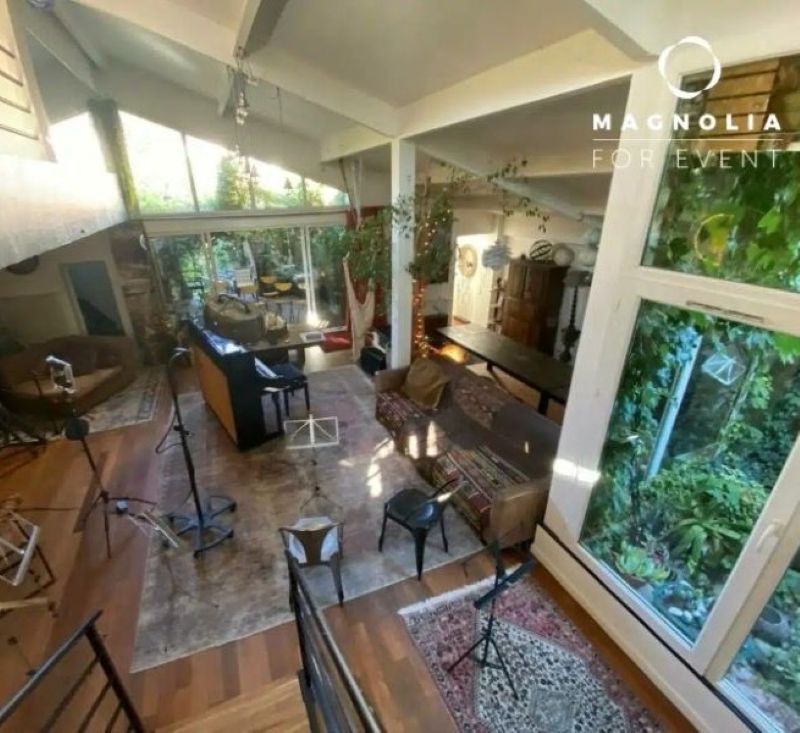 Chambre spacieuse et lumineuse à LE LOFT ART FEELING (93170) BAGNOLET avec parquet, grandes fenêtres, tapis, plantes et pupitres. La lumière naturelle remplit l'espace confortable avec de la verdure visible à l'extérieur. Le logo de Magnolia For Event est affiché dans le coin.