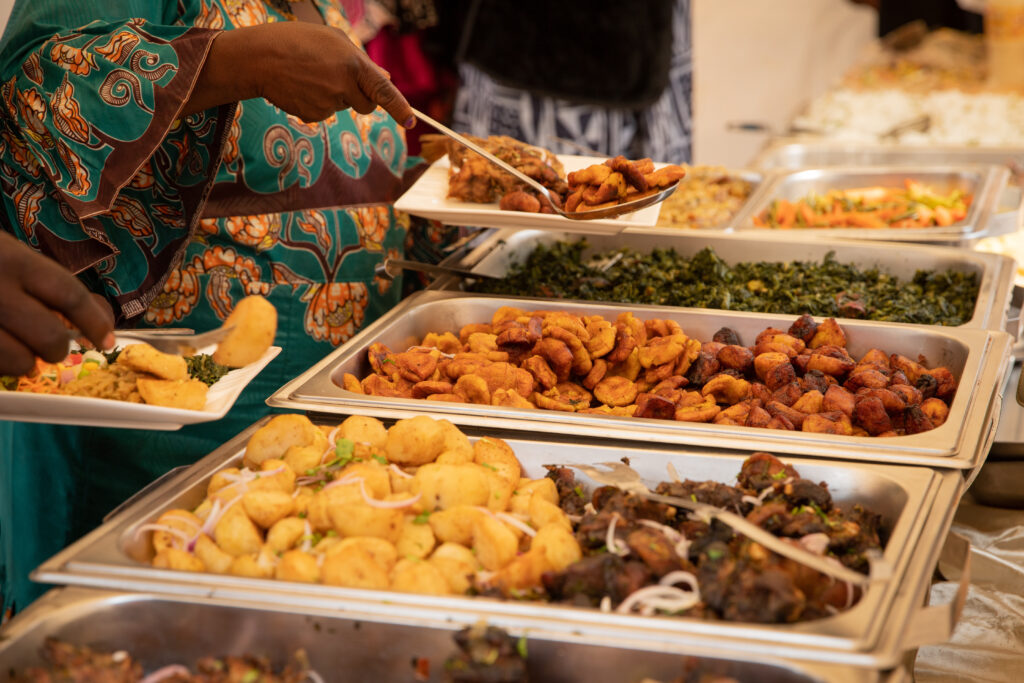 Une personne sert à manger lors d'un buffet composé de plateaux de bananes plantains frites, de légumes et de plats variés. Elle porte une tenue à motifs et tient une assiette blanche.
